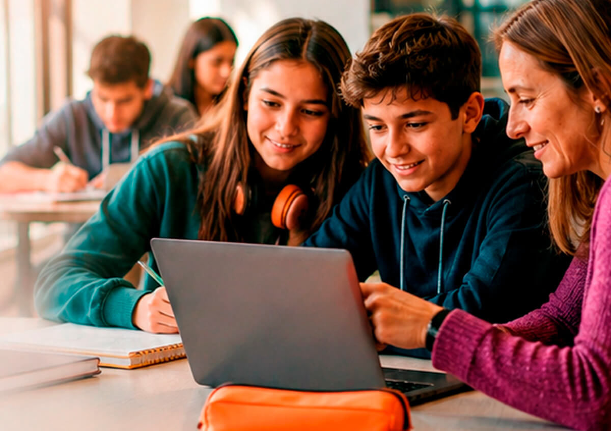 Docente acompañando a dos estudiantes que trabajan juntos en una notebook durante la clase